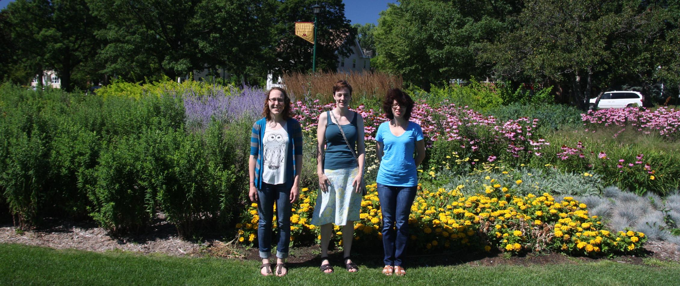 Three people standing outside in front of bushes and flowers