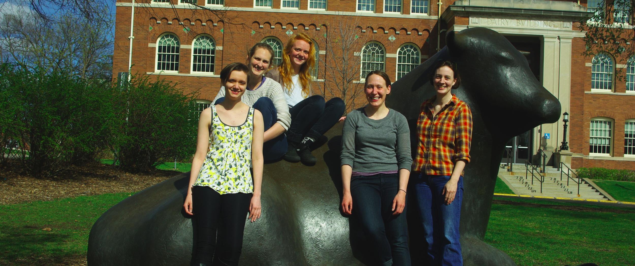 Five people standing next to or sitting on a large cow statue