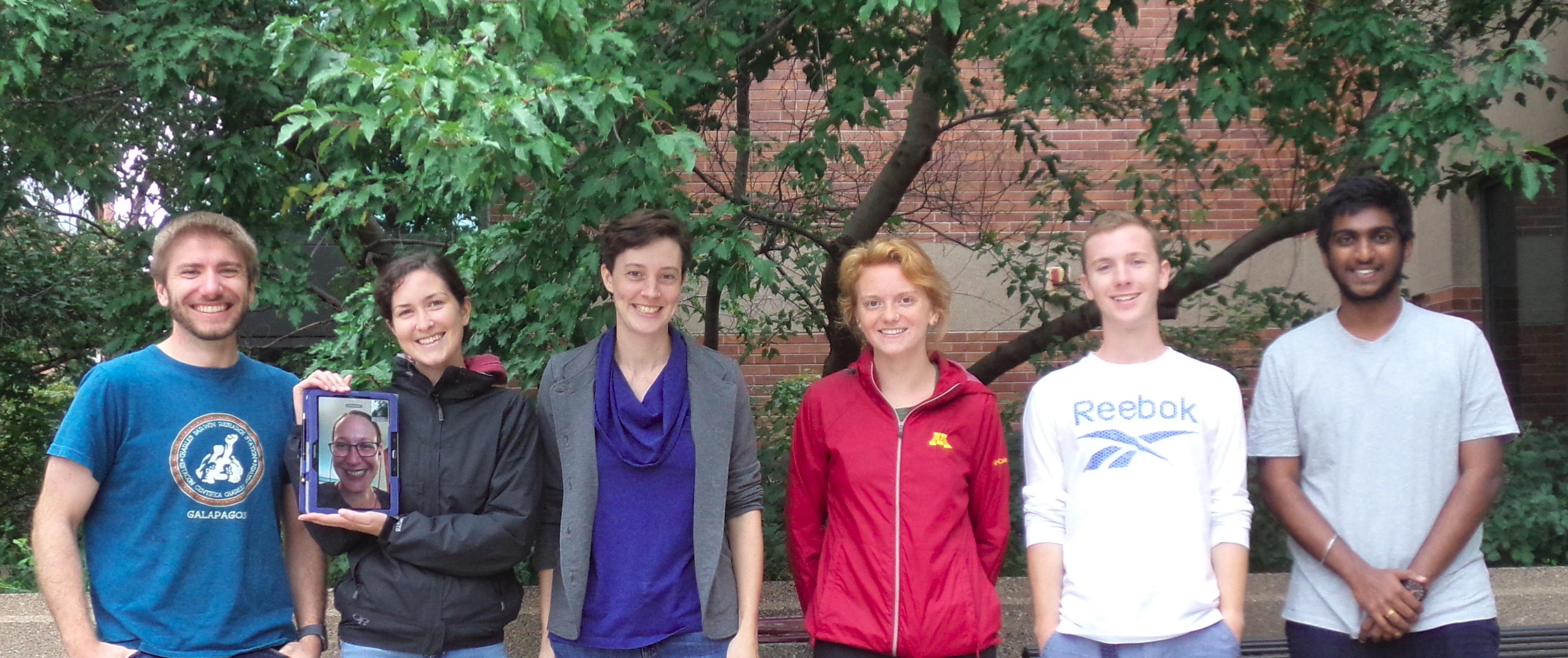 Six people standing with one holding an image of a seventh person, with a tree and building in the background