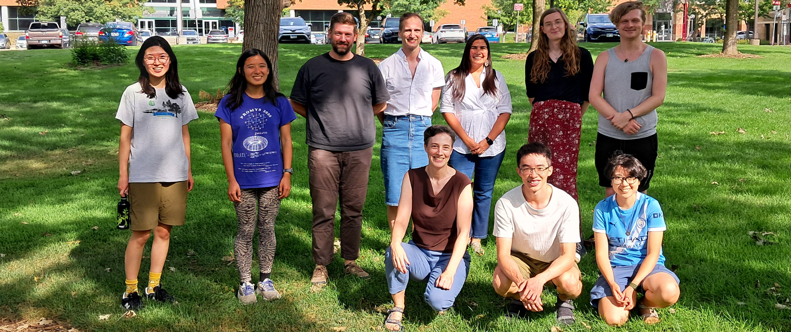 Ten people posing in front on a grassy lawn with tres in the background