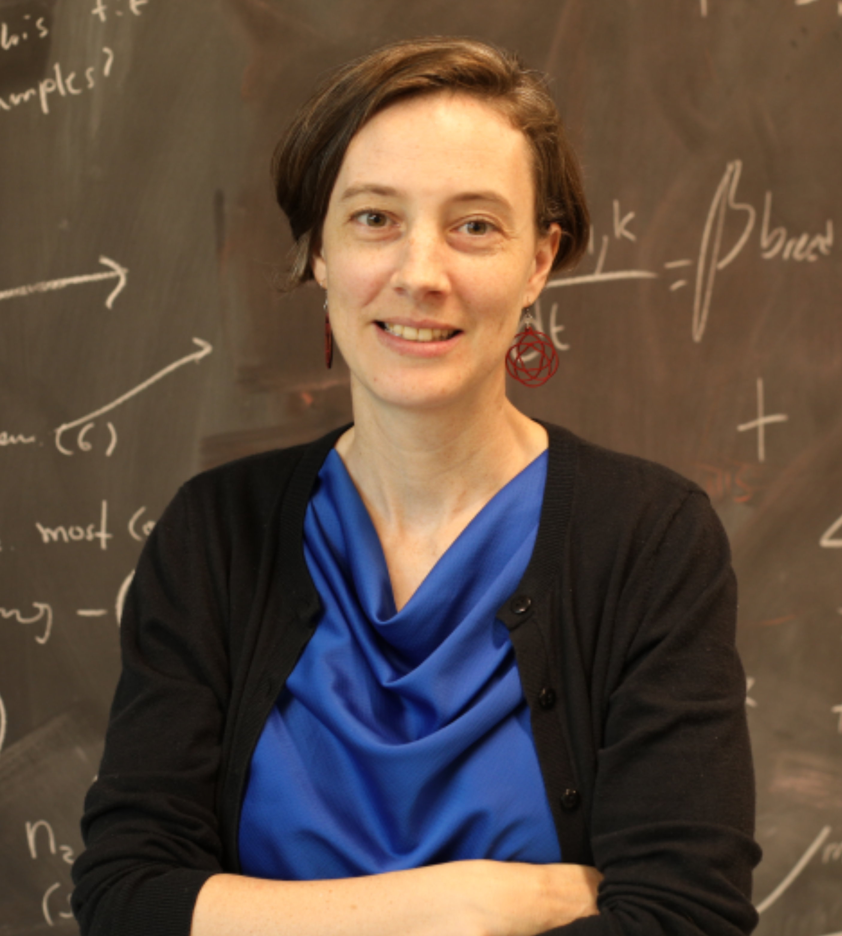 Portrait of Allison with short brown hear, wearing a blue top and black cardigan, standing in front of a chalk board