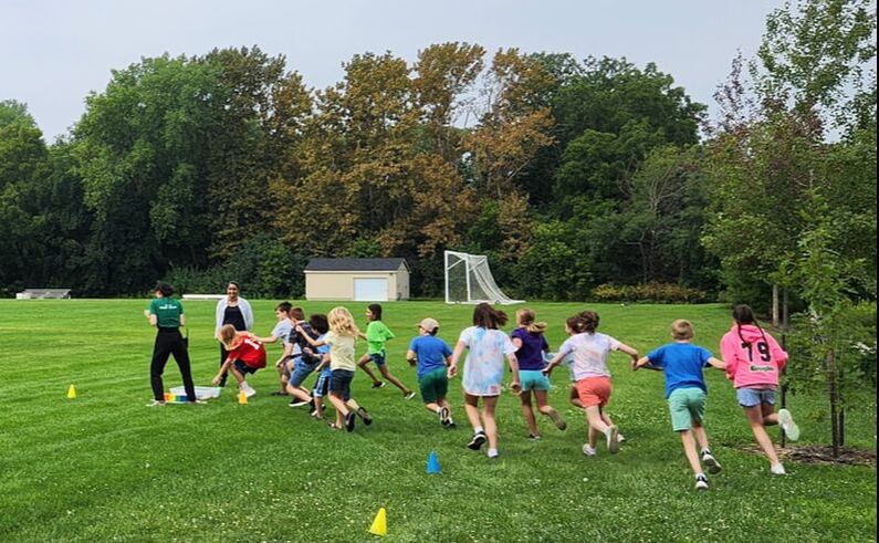 Children running in a grassy field next to a line of cones and towards a bucket on the ground